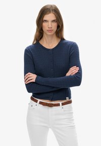 Granatowy bawełniany kardigan o warkoczowym splocie, okrągłym dekolcie i z zapięciem na guziki. Połączony z białymi spodniami i brązowym plecionym paskiem.