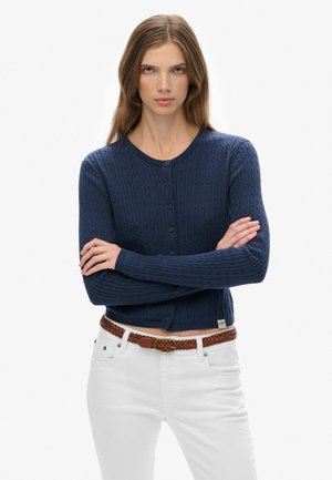 Cardigan di cotone blu navy con texture a trecce, scollo rotondo e chiusura con bottoni. Abbinato a pantaloni bianchi e una cintura intrecciata marrone.
