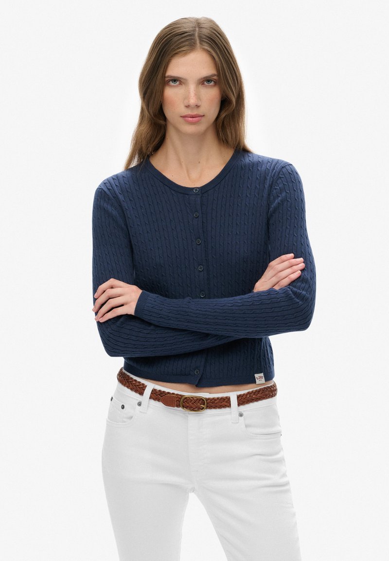 Granatowy bawełniany kardigan o warkoczowym splocie, okrągłym dekolcie i z zapięciem na guziki. Połączony z białymi spodniami i brązowym plecionym paskiem.