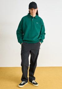 Groene lange mouw polo met twee knopen en geborduurd logo, gecombineerd met zwarte cargo broeken met ritszakken en zwarte sneakers.