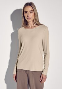 Beige pitkähihaine ribbitoppi pyöreällä kaula-aukolla, yhdistettynä ruskeisiin housuihin. Kankaan kuviointi on pehmeä ja se istuu hyvin.