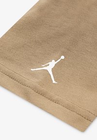 Béžová bavlněná tkanina má hladkou texturu s bílým logem Jumpman. Detailní záběr ukazuje švy podél okraje.