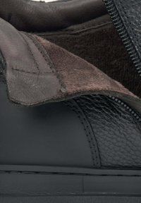 Wnętrze czarnego skórzanego buta z teksturowanym wzorem, ciemno brązową zamszową podszewką i detalem zamka, podkreślającym kontrastujące materiały.