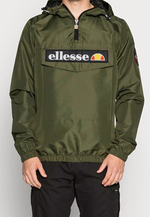 Mężczyzna w oliwkowo-zielonej wiatroodpornej kurtce z kapturem, z przednią kieszenią i logo "ellesse", w połączeniu z czarnymi spodniami.