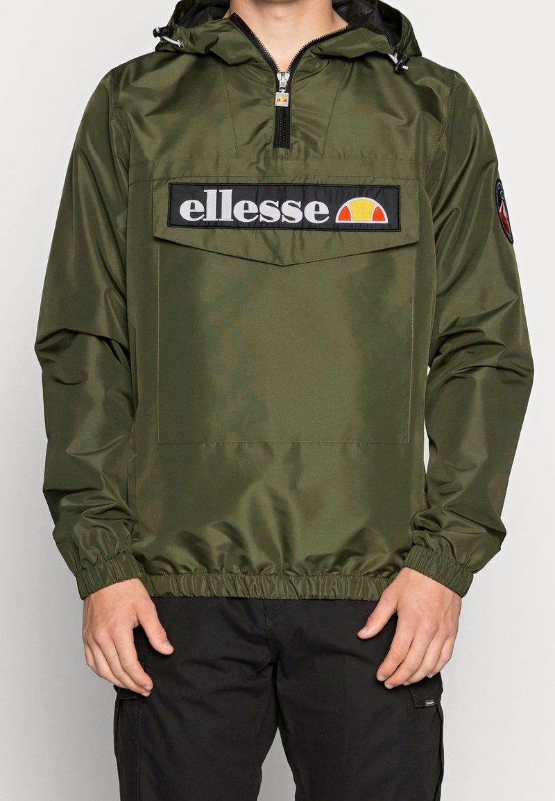 Mężczyzna w oliwkowo-zielonej wiatroodpornej kurtce z kapturem, z przednią kieszenią i logo "ellesse", w połączeniu z czarnymi spodniami.