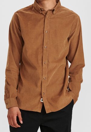 Mies, joka pukeutunut ruskeaan pitkähihainen nappipaitoihin samettikankaasta ja mustiin housuihin, toinen käsi taskussa, yksinkertaista vaaleaa taustaa vasten.