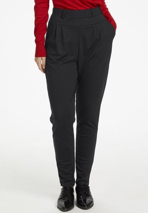 Personne portant un pantalon noir taille haute fuselé, des chaussures noires à lacets et un haut rouge à manches longues avec une main dans la poche.
