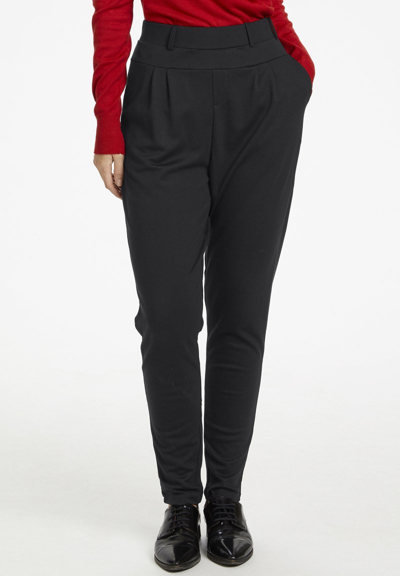 Personne portant un pantalon noir taille haute fuselé, des chaussures noires à lacets et un haut rouge à manches longues avec une main dans la poche.
