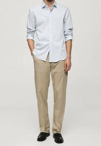 Camisa azul claro de manga larga con cuello abotonado y mangas remangadas, combinada con pantalones beige y zapatos negros sobre un fondo neutro.