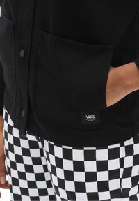 Giacca nera con bottoni sul davanti e tasche anteriori; abbinata a pantaloni con motivo a scacchi bianco e nero. Etichetta con logo Vans visibile sulla giacca.