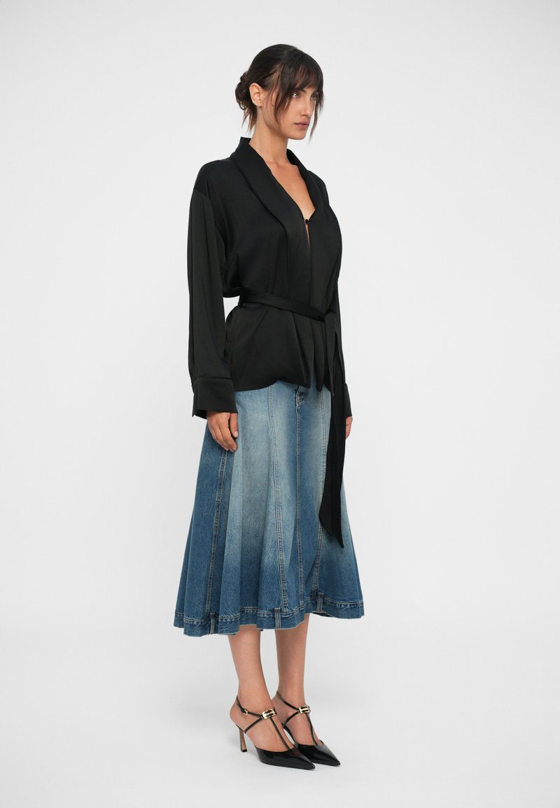 Schwarze Bluse mit Gürtel, kombiniert mit einem blauen Denim-Rock, der einen ausgestellten Schnitt und Saumdetails aufweist. Getragen mit schwarzen spitzen Absatzschuhen.