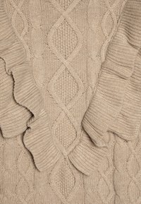 Beige kabelstrikket genser med et strukturert mønster med diamantformer og bølgede kanter langs halsen og ermene.