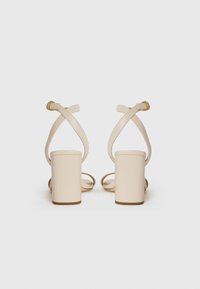 Beige heeled sandals with a block heel, cross-strap design, and adjustable ankle strap featuring a gold buckle detail. Transparent accents.