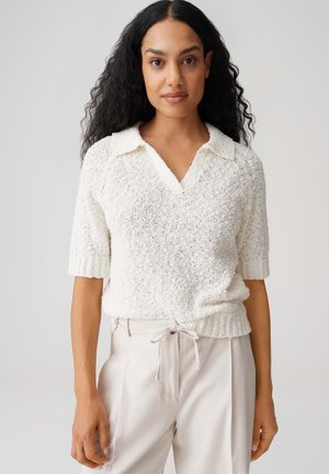 Vrouw met lang krullend haar, gekleed in een witte kortere mouwtrui met structuur en lichtbeige broek met een taillekoord.