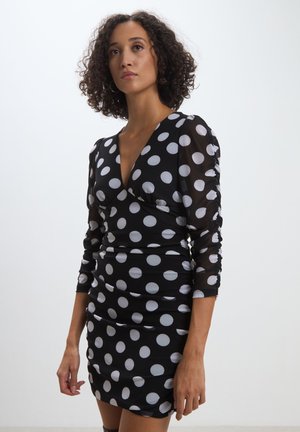 Abito nero con pois bianchi, caratterizzato da una profonda scollatura a V, maniche a tre quarti arricciate e una silhouette aderente. Tessuto liscio e elastico.