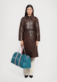 Donna con un cappotto di pelle marrone scuro con cintura, pantaloni crema e scarpe nere, che tiene un borsone verde acqua con manici a righe e testo in corsivo.