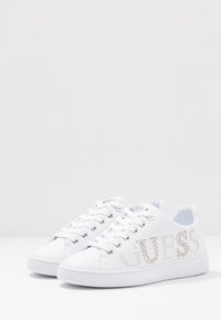 Baskets blanches en matériau synthétique, avec une semelle texturée, un bout rond et un logo orné de strass sur le côté.