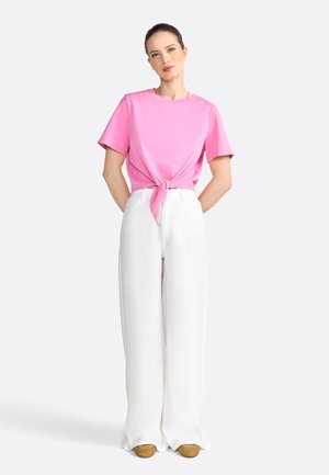 Roze katoenen T-shirt geknoopt in de taille, gecombineerd met witte wijde broek en bruine schoenen, tegen een neutrale achtergrond.