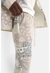 Pantaloni cargo beige e bianchi con un motivo che presenta vari disegni paisley e floreali, dotati di tasche e vita con cordoncino.