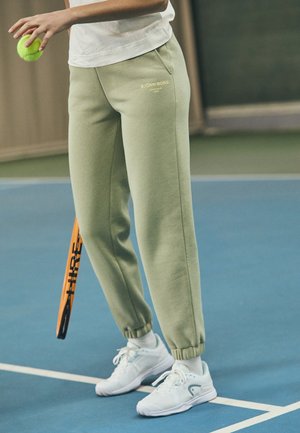 Lichtgroene sweatpants met geribde boorden hebben een gouden logo. Gepaard met witte sportschoenen, een tennisbal en een racket op een blauwe tennisbaan.