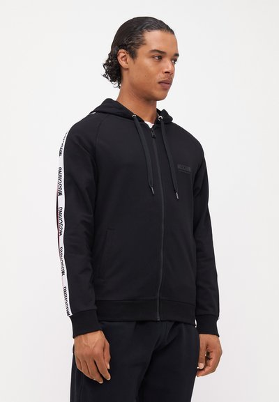 Črna zip-up kapuca iz mehkega materiala, ki ima sprednji žep, kapuco na vrvico in bele stranske črte z logotipom blagovne znamke.