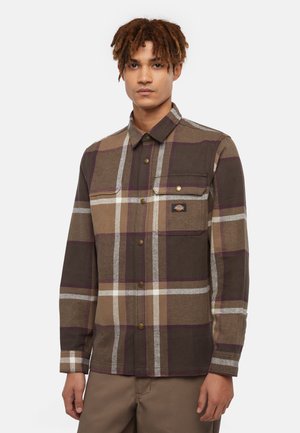 Camisa de franela a cuadros marrón con rayas beige y blancas, que presenta un cuello abotonado, bolsillo en el pecho y un acento de logo metálico.