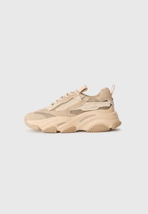 Beige sneaker made of mesh and suede, featuring a thick sole with textured patterns, lace-up design, and a looped back pull tab.