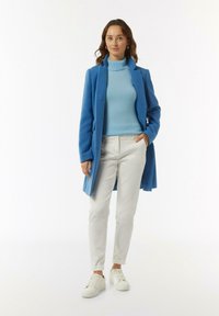 Lichtblauwe jas, bijpassende coltrui, witte broek en witte sneakers. Glad materiaal met een getailleerde pasvorm en knoopsluiting aan de voorkant.