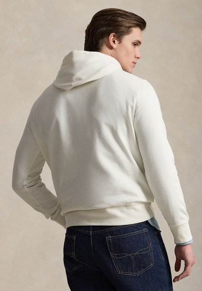 Νέος άνδρας σε προφίλ, φοράει λευκή φούτερ με κουκούλα και σκούρο μπλε τζιν, μπροστά σε απλό μπεζ φόντο.