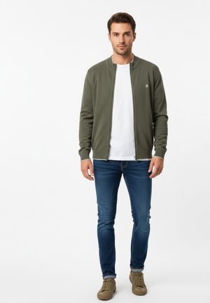 Hombre de pie con chaqueta verde oliva con cremallera, camiseta blanca, vaqueros azules y zapatillas marrones sobre un fondo blanco.