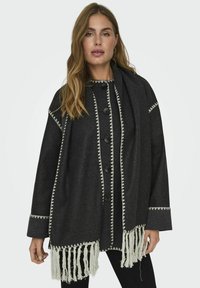 Cappotto di lana grigio scuro con maniche lunghe, grandi bottoni neri, cuciture a zigzag bianche e dettaglio di orlo sfrangiato.