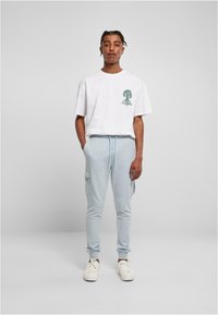 Hvit t-skjorte med kort erme og grønt grafisk logo, sammen med lyseblå joggebukser med cargo-lommer og elastiske mansjetter. Hvite sneakers.