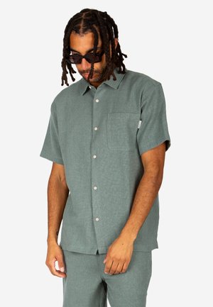 Mann mit Dreadlocks und Sonnenbrille, der ein grünes kurzärmeliges Hemd mit Knöpfen und passende Hose trägt, steht vor einem weißen Hintergrund.