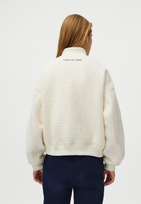 Kermakoska fleece-paita, jossa on korkea kaulus, ylipitkä malli ja ribbireunat. Takana on huomaamaton logo.