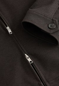 Poignet de veste en tissu marron foncé avec bouton et deux tirettes de fermeture éclair en métal argenté sur la surface du tissu.