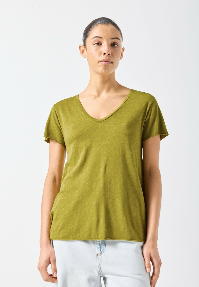 Jeune femme aux cheveux tirés en arrière portant un T-shirt olive à col en V et un jean bleu clair sur un fond blanc uni.