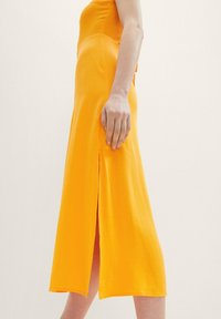 Gelbes ärmelloses Kleid mit enganliegendem Oberteil und einem seitlichen Schlitz. Hergestellt aus strukturiertem Stoff, reicht es bis zur Mitte der Wade und betont eine glatte Silhouette.