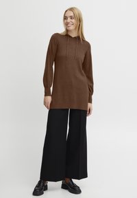 Sweat à capuche marron avec des manches longues et des bords côtelés, associé à un large pantalon noir et des mocassins noirs. Texture lisse, design décontracté.