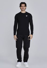 Melna garenagalvu krekls ar mazu baltu logo uz krūtīm, apvienots ar melniem cargo biksēm, kurām ir kabatas un zelta akcenti.