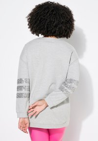 Felpa grigia con maniche lunghe caratterizzata da dettagli a righe di paillettes sulle maniche. Tessuto morbido e vestibilità rilassata, indossata con leggings rosa.