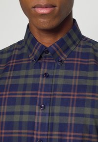Camicia con colletto in un motivo a quadretti blu navy, verde e marrone. Presenta un colletto e una chiusura frontale con bottoni scuri. Tessuto in cotone.