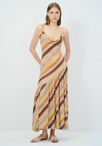 Maxikleid mit diagonalen Streifen in Braun, Gelb und Orange; Wasserfall-Ausschnitt, dünne Träger und ein geraffter Rockbereich. Getragen mit beigen Ballerinas.