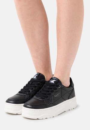 Jambes portant des baskets plateformes en cuir noir à lacets avec des semelles blanches épaisses sur un fond clair uni.