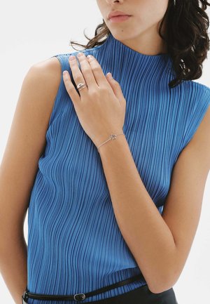 Top sin mangas plisado azul con un cuello alto. Anillo plateado en una mano y pulsera de estrella plateada en la muñeca. Cinturón negro como acento.