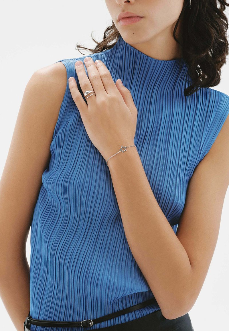 Top sin mangas plisado azul con un cuello alto. Anillo plateado en una mano y pulsera de estrella plateada en la muñeca. Cinturón negro como acento.