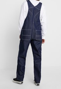 Tummansininen farkkukokonaisuus, jossa säädettävät olkaimet, kaksi suurta etutaskua ja kontrastina valkoiset saumanompeleet.