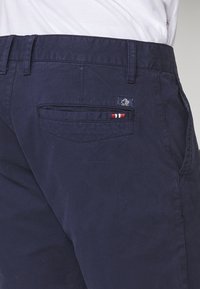 Donkerblauwe broek met een achterzak, voorzien van een klein stikte detail in rood, wit en blauw, en een label met een esdoornblad.