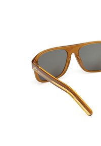 Ochelari de soare din plastic transparent marron cu lentile gri și un accent în formă de frunză pe ramă. Temoli drepți care se subțiază spre capete.