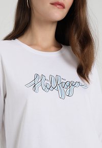 T-shirt bianco a maniche lunghe con logo a righe blu e nere sul davanti. Realizzato in tessuto morbido con colletto rotondo.