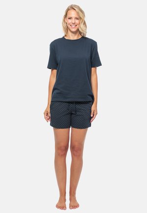 T-shirt di cotone navy a maniche corte abbinato a pantaloni corti neri con pois bianchi. Il modello è in piedi a piedi nudi su uno sfondo neutro.